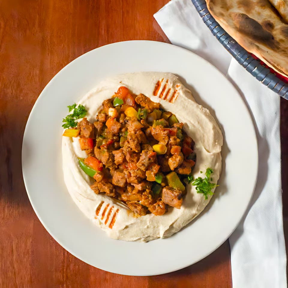 Hummus topped with seasoned meat and colorful vegetables, garnished with parsley, served with flatbread.