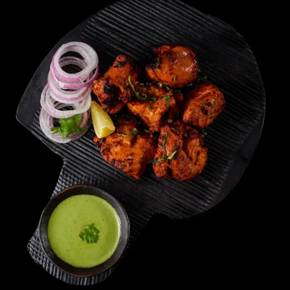 Grilled chicken tikka with red onion, lemon, cilantro, and green chutney on a black platter.